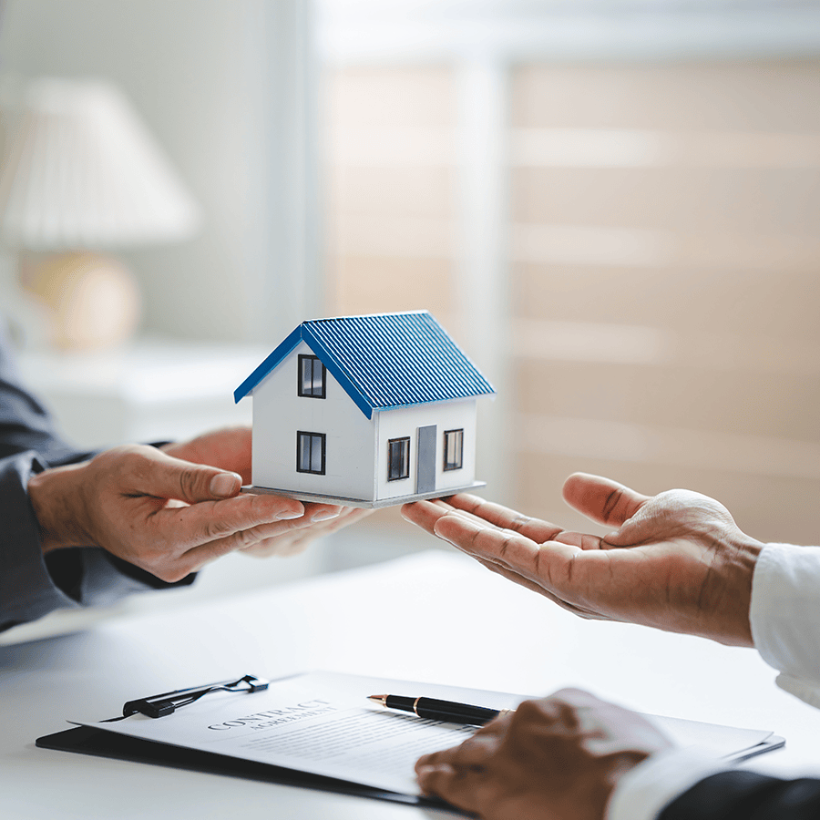 Zwei Hände übergeben ein Modellhaus mit blauem Dach, während ein Vertrag auf einem Tisch liegt.