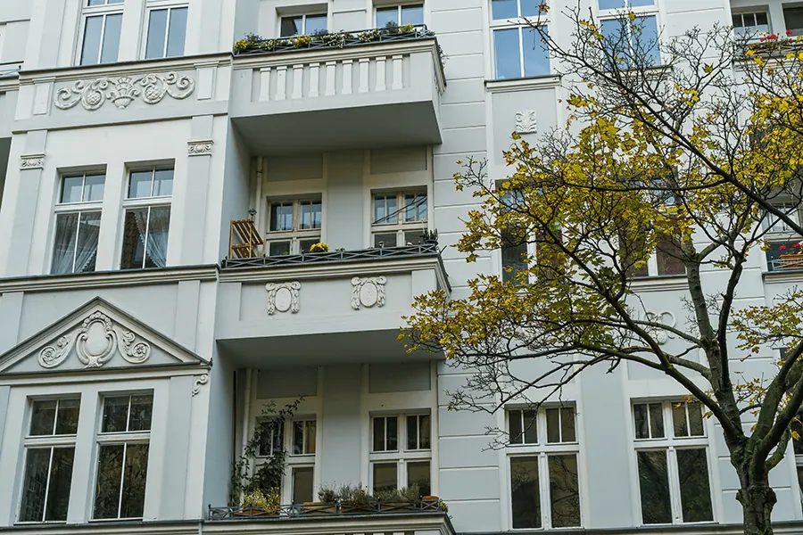 Mehrfamilienhaus mit hellblauer Fassade, Balkonen und dekorativen Elementen, umgeben von einem Baum mit gelben Blättern.